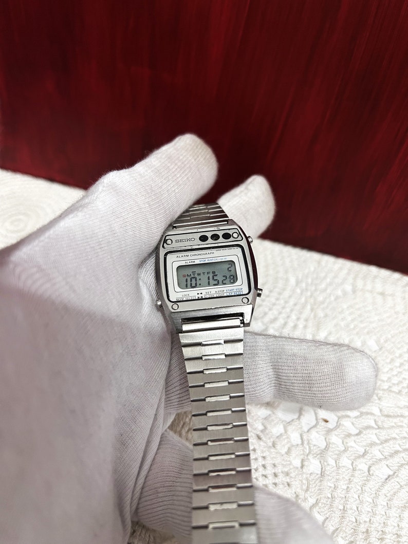 Puede incluir: Un reloj digital Seiko plateado con una correa de metal plateado. La esfera del reloj muestra la hora, la fecha y el d&iacute;a de la semana. El reloj tiene una pantalla digital negra con n&uacute;meros blancos. El reloj tiene un bot&oacute;n negro en la parte superior derecha de la esfera.