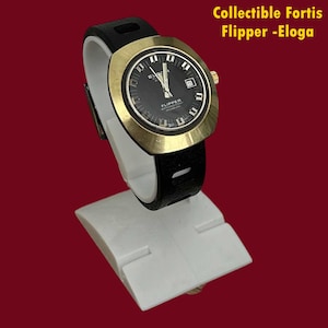 Puede incluir: Un reloj de pulsera vintage Fortis Flipper Eloga con caja redondeada en tono dorado, esfera negra con marcadores de hora blancos y correa negra. La esfera muestra el texto FLIPPER WATERRESISTANT INCABLOC. El texto Collectible Fortis Flipper -Eloga es visible.