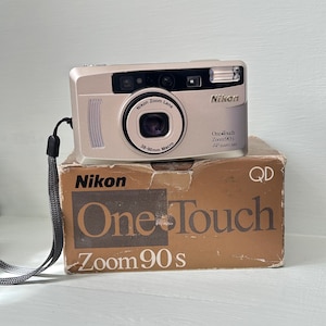 Könnte beinhalten: Eine silberne Nikon One Touch Zoom 90s Kamera mit einem 38-90mm Makroobjektiv liegt auf der Originalverpackung. Die braune Schachtel trägt das Nikon-Logo und den Schriftzug "One Touch Zoom 90s". Ein schwarzer Gurt ist an der Kamera befestigt.