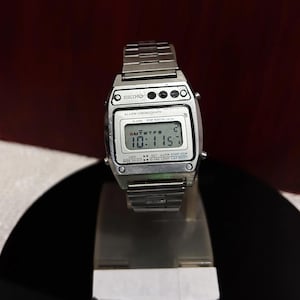 Puede incluir: Un reloj digital Seiko plateado con una esfera negra y una correa de metal plateado. El reloj tiene una pantalla digital que muestra la hora, la fecha y el d&iacute;a de la semana. El reloj tambi&eacute;n tiene una funci&oacute;n de cron&oacute;metro, alarma y luz.