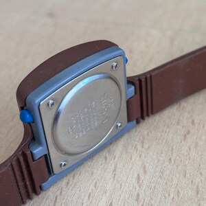 May include: Close-up of a vintage brown and blue digital watch. The watch has a rectangular face with a brown case and a blue button. The watch band is brown. The back of the watch is silver.