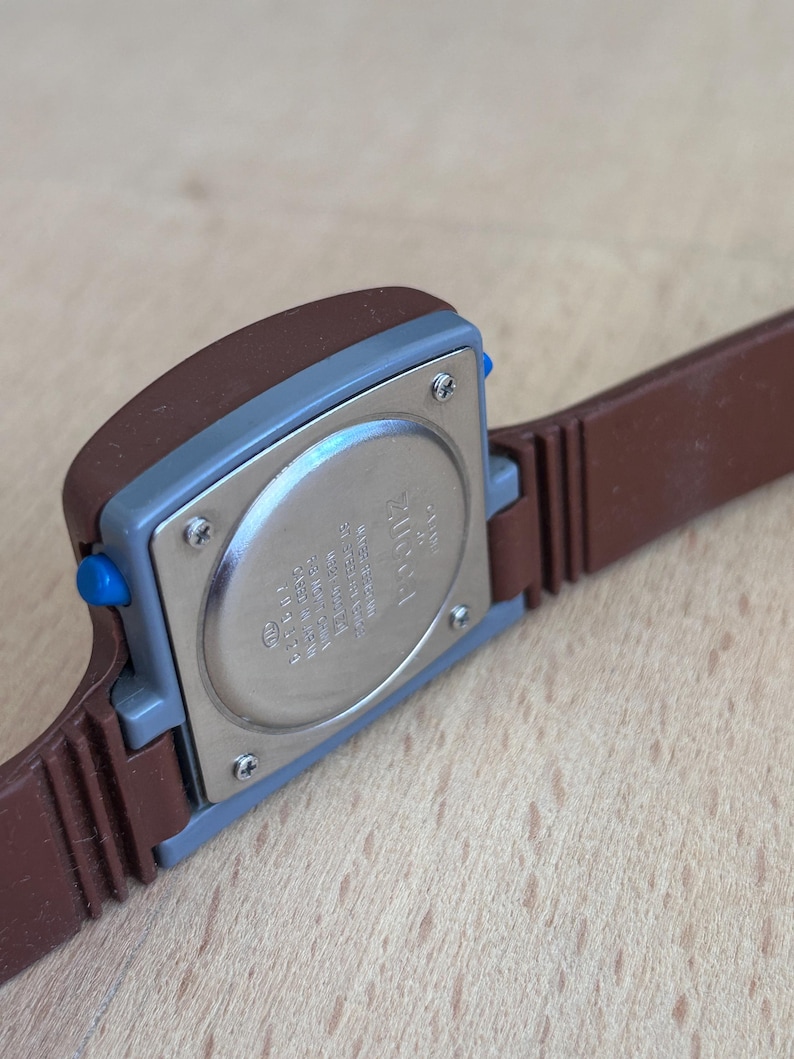 May include: Close-up of a vintage brown and blue digital watch. The watch has a rectangular face with a brown case and a blue button. The watch band is brown. The back of the watch is silver.