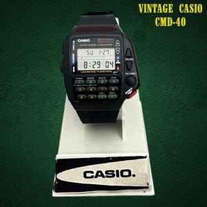 May include: A black Casio CMD-40 vintage digital wristwatch featuring a built-in calculator and TV remote control function. The watch face displays the time and date, with a numeric keypad below the screen and dedicated buttons for TV volume and power.