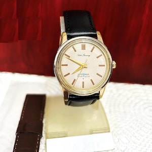 May include: A gold-tone Seiko Timeway wristwatch with a white face and black leather band. The watch face has black Roman numeral hour markers and a black second hand. The watch is water-resistant.