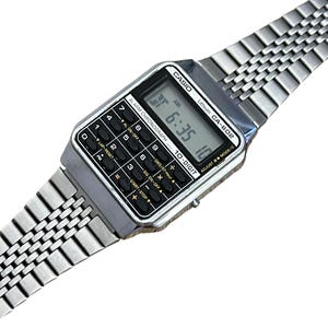 May include: A silver Casio CA-602 digital watch with a calculator function. The watch displays the time as 6:35 AM. The watch has a rectangular face with black buttons and a silver link band. The watch is a vintage style.