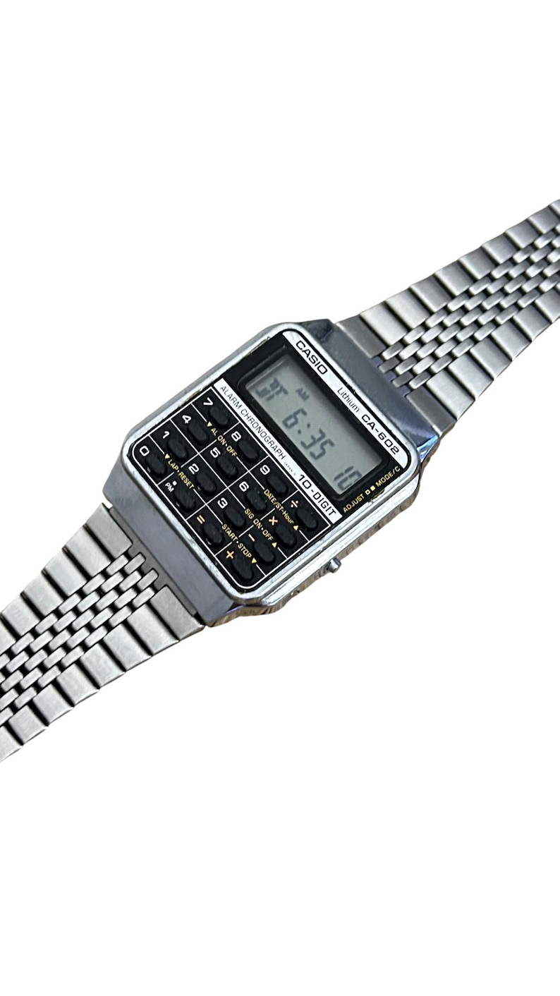 May include: A silver Casio CA-602 digital watch with a calculator function. The watch displays the time as 6:35 AM. The watch has a rectangular face with black buttons and a silver link band. The watch is a vintage style.