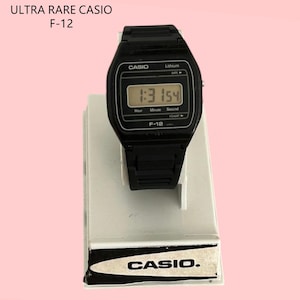 May include: A black Casio F-12 digital watch with a rectangular face displaying the time. The watch is on a white display stand with the Casio logo. The text "ULTRA RARE CASIO F-12" is at the top.