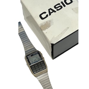 May include: A vintage Casio calculator watch with a silver-tone metal band and a digital display showing the time. The watch has a calculator keypad and is next to a white box with the brand name "CASIO" printed in black.