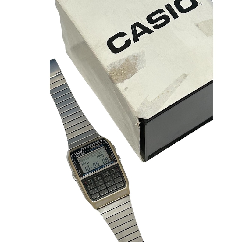 May include: A vintage Casio calculator watch with a silver-tone metal band and a digital display showing the time. The watch has a calculator keypad and is next to a white box with the brand name "CASIO" printed in black.