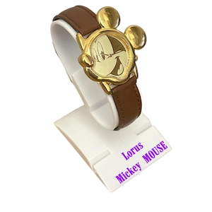 May include: A gold-toned Mickey Mouse watch with a brown leather band. The watch face features Mickey's face, and the watch is displayed on a white stand. The text "Lorus Mickey Mouse" is visible on the stand.