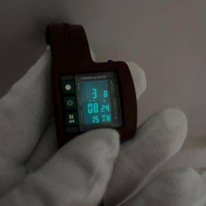 May include: A brown digital watch with a rectangular face displaying the time and date in a teal color. The watch has a matching brown strap and is held by a gloved hand. The watch face reads "CABANE de ZUCCA".