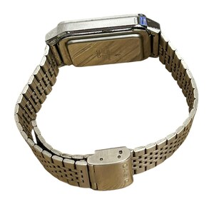 May include: A silver-toned digital watch with a rectangular face and a metal band. The watch band is made of small, interconnected links. The clasp is closed. The watch is angled, showing the back of the watch face.