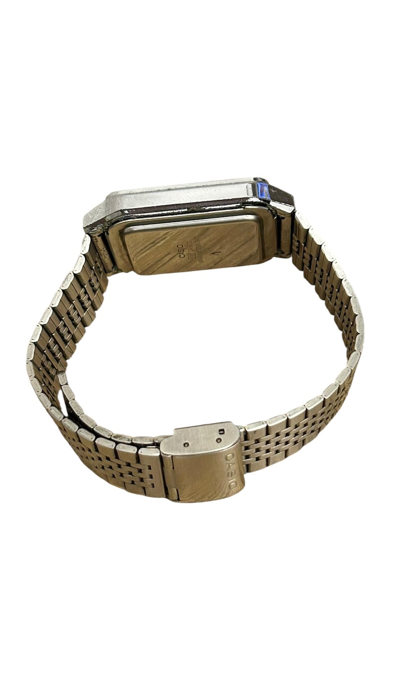 May include: A silver-toned digital watch with a rectangular face and a metal band. The watch band is made of small, interconnected links. The clasp is closed. The watch is angled, showing the back of the watch face.