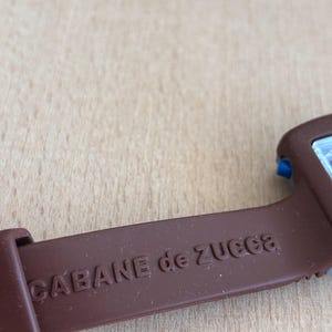 May include: Close-up of a brown watch strap with the text "CABANE de ZUCCA" embossed on it. The strap is made of a flexible material and is attached to a watch face with blue accents.