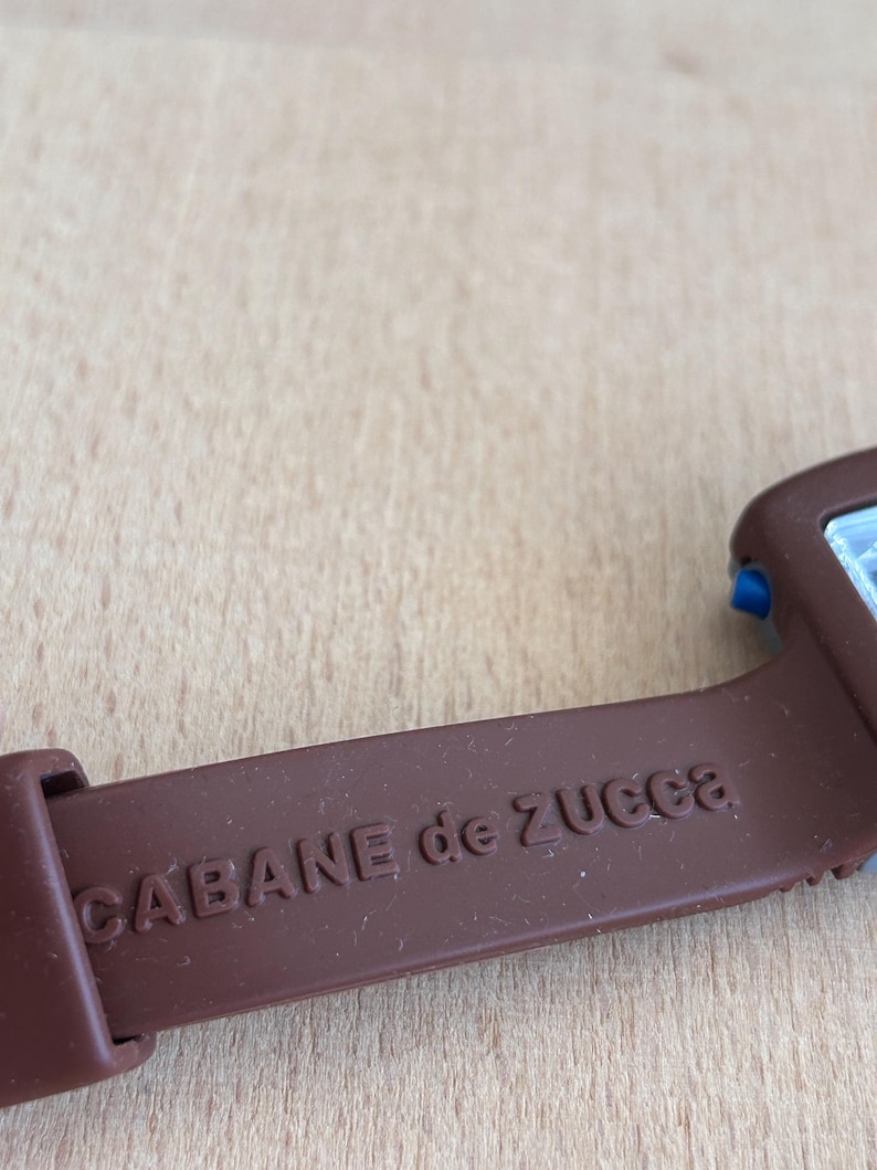 May include: Close-up of a brown watch strap with the text "CABANE de ZUCCA" embossed on it. The strap is made of a flexible material and is attached to a watch face with blue accents.
