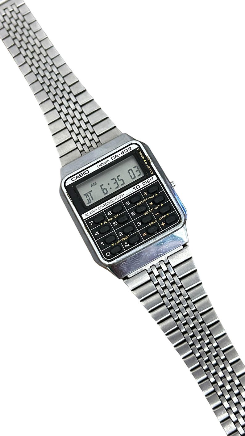 May include: A vintage Casio CA-602 digital watch with a silver-tone metal band. The watch face displays the time as 6:35:03 AM. It features a calculator keypad and the text "ALARM CHRONOGRAPH".