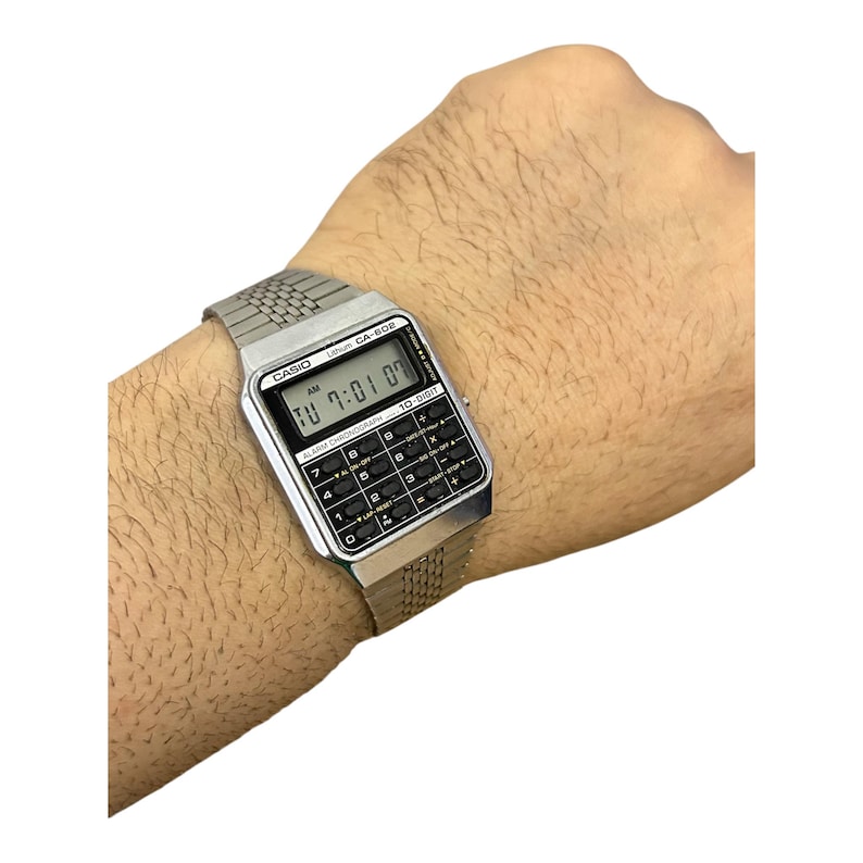 May include: Silver-toned Casio CA-506 calculator watch with a black keypad and digital display showing the time. The watch has a mesh band and is worn on a wrist.