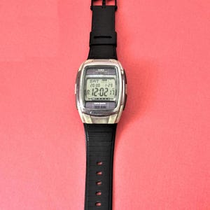 Puede incluir: Un reloj Casio Data Bank plateado con una correa negra. La esfera del reloj muestra la fecha, el día de la semana y la hora. El reloj está configurado a las 12:02:17.