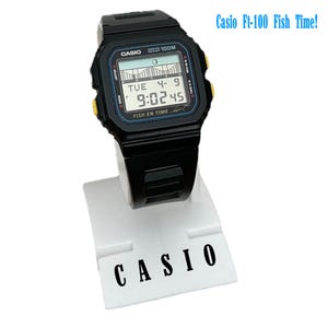 May include: Black Casio Ft-100 digital watch with a black band, displayed on a white stand. The watch face shows the time, date, and a graphic. The text "Casio Ft-100 Fish Time!" is visible above the watch.