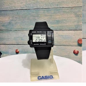 以下が含まれることがあります： 黒いバンドが付いた黒いカシオデジタル腕時計。時計の文字盤には、時間、日付、メモが表示されています。時計は、カシオのロゴが付いた白いディスプレイスタンドの上に置かれています。