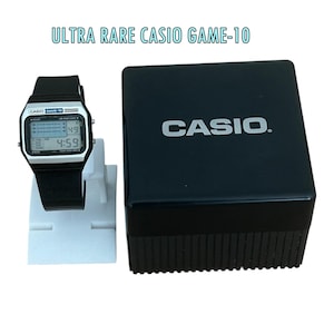May include: A black Casio Game-10 digital watch with a silver face and black strap, displayed next to a black Casio watch box. The watch screen shows the time 4:59 and the text ULTRA RARE CASIO GAME-10 is visible above the watch and box.