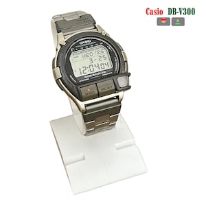 Puede incluir: Un reloj digital Casio DB-V300 plateado con bisel negro y pantalla digital que muestra la fecha y la hora. El reloj tiene botones etiquetados "REC" y "PLAY". El reloj se muestra en un soporte blanco.