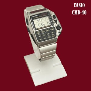May include: A silver Casio CMD-40 digital watch with calculator and remote control features. The watch shows the time and date on a digital display. The watch is displayed on a white stand against a maroon background.