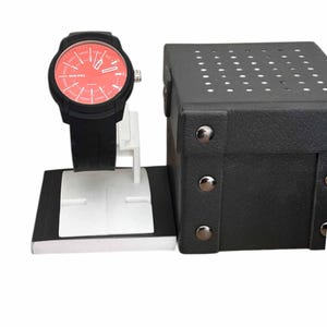 May include: Black wristwatch with a red dial, displayed on a white and black stand. The watch has a black strap and the word "DIESEL" is printed on the dial. A black box with silver rivets is next to the watch.