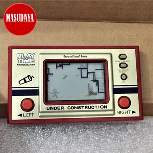 May include: A red and gold handheld video game console with a black and white screen. The screen displays a pixelated image of a construction site with a character running. The console has buttons labeled "LEFT", "RIGHT", "GAME1", "GAME2", "TIME ALARM", "TIME SET", and "ACL". The console is labeled "PLAY TIME MASUDAYA" and "Crystal Head Game". The text "UNDER CONSTRUCTION" is displayed on the screen.