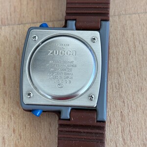May include: A close-up of a Cabane de Zucca watch. The watch has a gray square face with blue buttons and a brown strap. The back of the watch has text that reads "WATER RESISTANT ST. STEEL + PLASTICS W621-4000 [20] FB MOV'T CHINA GASED IN JAPAN 709329".