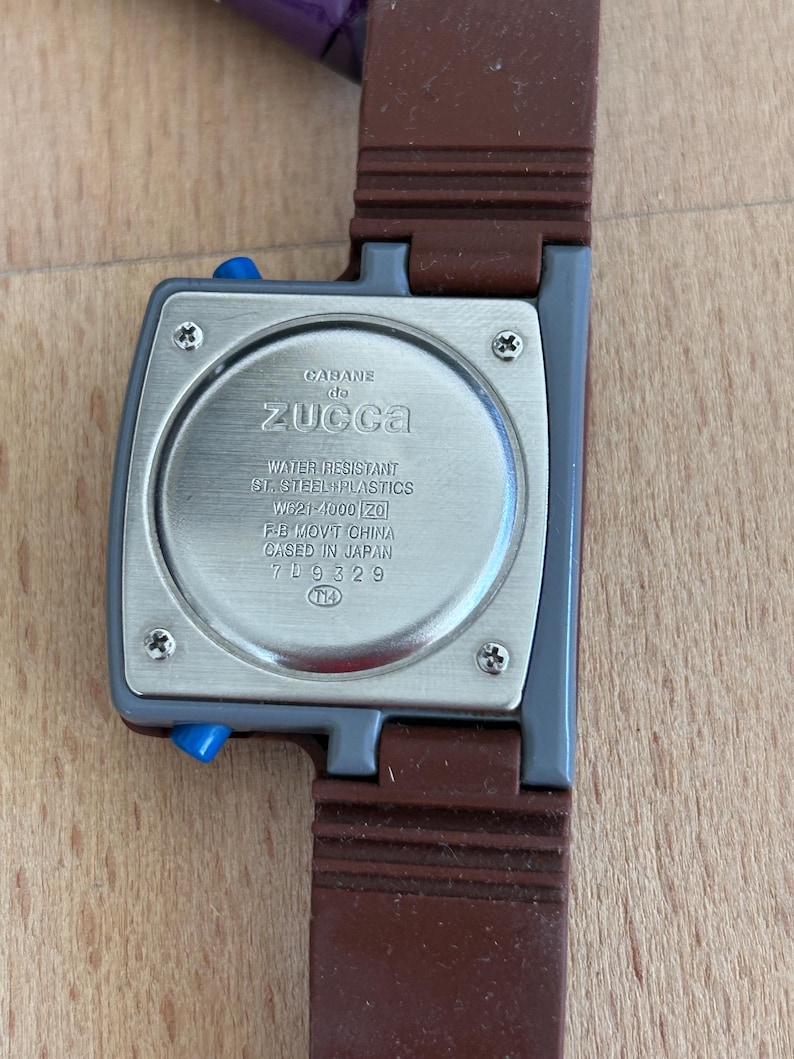 May include: A close-up of a Cabane de Zucca watch. The watch has a gray square face with blue buttons and a brown strap. The back of the watch has text that reads "WATER RESISTANT ST. STEEL + PLASTICS W621-4000 [20] FB MOV'T CHINA GASED IN JAPAN 709329".