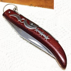 May include: A folding pocket knife with a dark red wooden handle and a silver blade. The handle features decorative silver accents and the word "OKAPI". A silver ring is attached to the end of the handle.