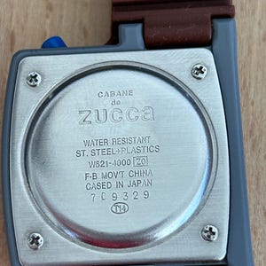 May include: Close-up of a Cabane de Zucca watch. The watch has a rectangular face with a silver-colored back and a brown strap. The back of the watch has the text "WATER RESISTANT ST. STEEL+PLASTICS" and other details.