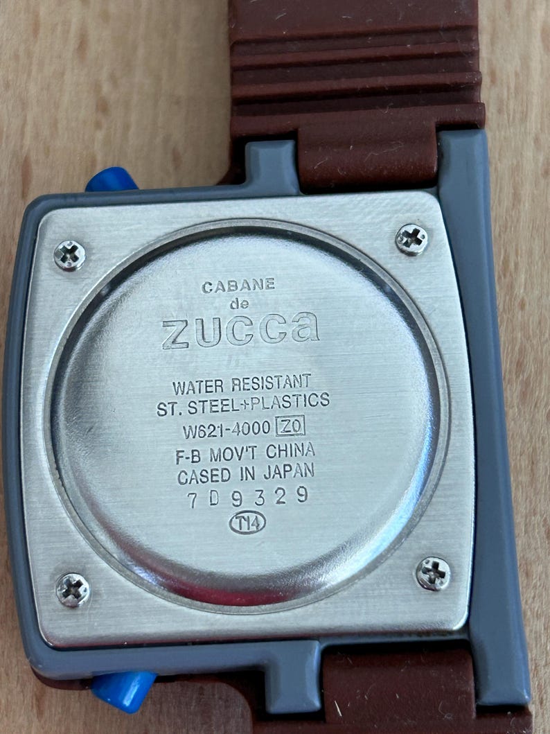 May include: Close-up of a Cabane de Zucca watch. The watch has a rectangular face with a silver-colored back and a brown strap. The back of the watch has the text "WATER RESISTANT ST. STEEL+PLASTICS" and other details.