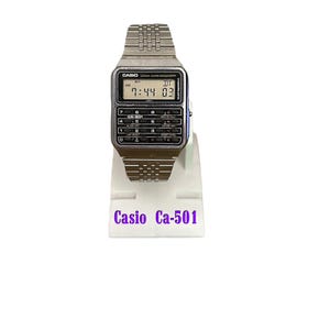 May include: A silver Casio Ca-501 digital watch with a calculator keypad. The watch displays the time as 7:44 and the date as 03. The watch is on a white stand with the words "Casio Ca-501" in purple.