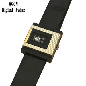 May include: A vintage Agon Digital Swiss watch with a black strap and a gold-toned rectangular face. The watch face displays the time and the brand name. The watch has a classic, retro design.