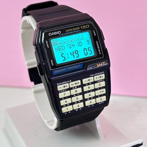 Puede incluir: Un reloj digital Casio Data Bank 150 negro con una calculadora. La esfera del reloj muestra la hora, la fecha y el día de la semana. La calculadora tiene una variedad de funciones, incluyendo suma, resta, multiplicación, división y memoria.