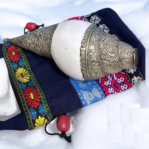 Puede incluir: Una concha blanca con adornos plateados, descansando sobre una bolsa de tela azul y roja con un borla roja.