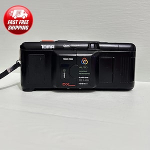 Puede incluir: Una cámara de película negra Toma DX-L de 35 mm con una lente roja y una lente de enfoque libre. La cámara tiene un flash incorporado y una función de rebobinado automático. La cámara está hecha en Japón.