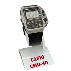 May include: A vintage Casio CMD-40 watch with calculator and remote control features. The watch has a grey body, black strap, and a digital display showing the time and date. The watch is displayed on a white stand with the text "CASIO CMD-40".