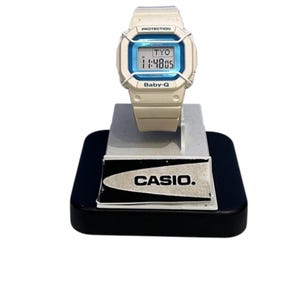 May include: A white Casio Baby-G digital watch with a blue display. The watch is on a white stand with the Casio logo.