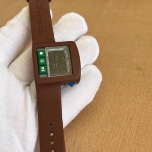 May include: A brown Cabane de Zucca digital watch. The watch has a rectangular face with a digital display showing the time and date. The watch band is brown and has the brand name printed on it. The watch is held by a gloved hand.