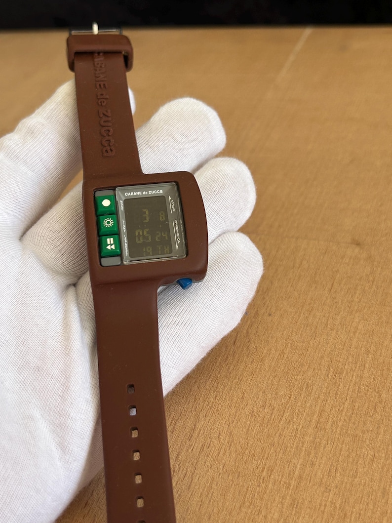 May include: A brown Cabane de Zucca digital watch. The watch has a rectangular face with a digital display showing the time and date. The watch band is brown and has the brand name printed on it. The watch is held by a gloved hand.