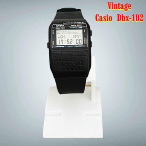 Puede incluir: Un reloj digital Casio DBX-102 negro vintage con un diseño de teclado de calculadora. El reloj muestra la hora y otros datos en una pantalla digital. El reloj está sobre un soporte blanco. El texto "Vintage Casio Dbx-102" está en la esquina superior derecha.
