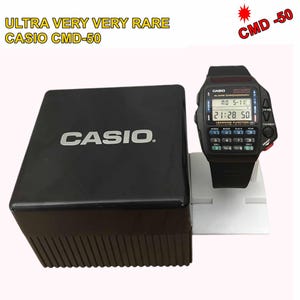 Op de afbeelding: Een zwarte Casio CMD-50 horloge met een rekenmachinetoetsenbord en digitale display, naast de zwarte doos. Het horloge toont de tijd en heeft de tekst "CASIO" op de wijzerplaat. De doos heeft het woord "CASIO" in het wit.