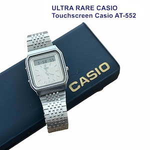 Könnte beinhalten: Eine silberfarbene Casio AT-552 Touchscreen-Uhr mit rechteckigem Zifferblatt, analogen Zeigern und einer Digitalanzeige, die 3:58 55 anzeigt. Die Uhr hat ein Metallgliederarmband und liegt auf einer dunkelblauen Box mit dem goldenen Schriftzug CASIO.