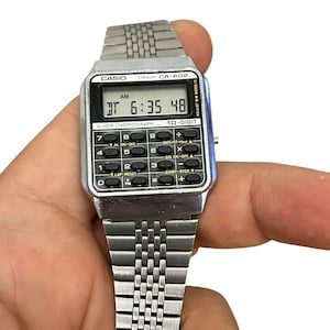 May include: A silver Casio CA-602 digital watch with a rectangular face displaying the time 6:35:48 AM. The watch has a metal band and multiple function buttons. The watch is held in a hand.