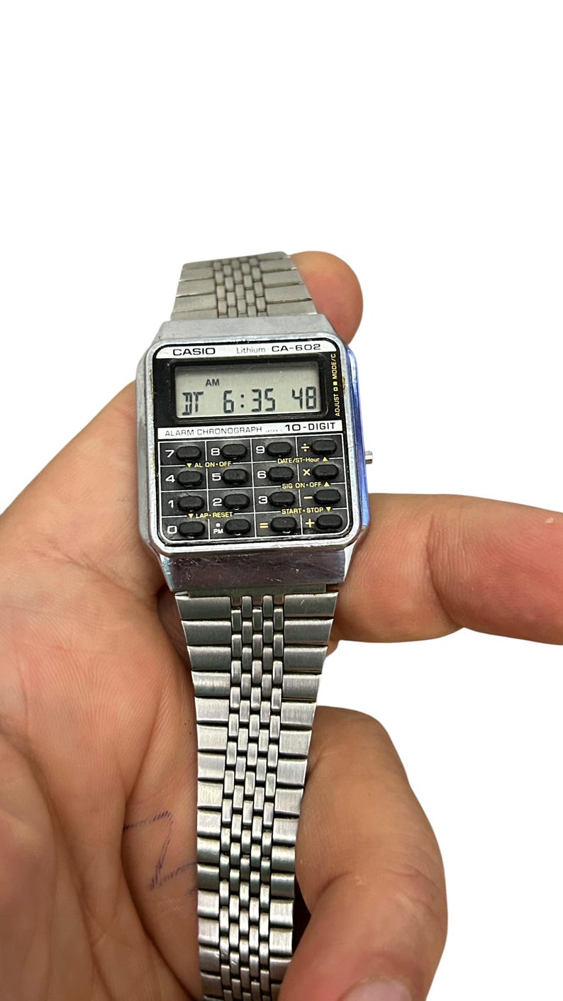May include: A silver Casio CA-602 digital watch with a rectangular face displaying the time 6:35:48 AM. The watch has a metal band and multiple function buttons. The watch is held in a hand.