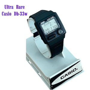 Puede incluir: Un reloj digital Casio Db-33w negro con una esfera rectangular y una correa negra. El reloj muestra la hora y la fecha. El texto "Ultra Rare Casio Db-33w" está encima del reloj. El reloj está sobre un soporte de exhibición blanco con el logotipo de Casio.