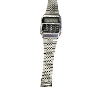 May include: Silver-toned Casio CA-502 calculator watch. The watch features a rectangular digital display showing the time, with a full calculator keypad below. The watch band is made of small, connected metal links.